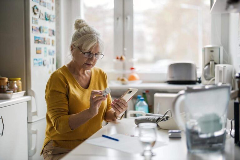 A person reading a prescription bottle label at their kitchen table with medication bottles nearby.