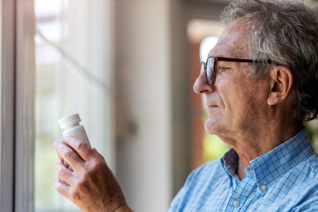 A person using a weekly pill organizer at a kitchen table with a glass of water nearby.